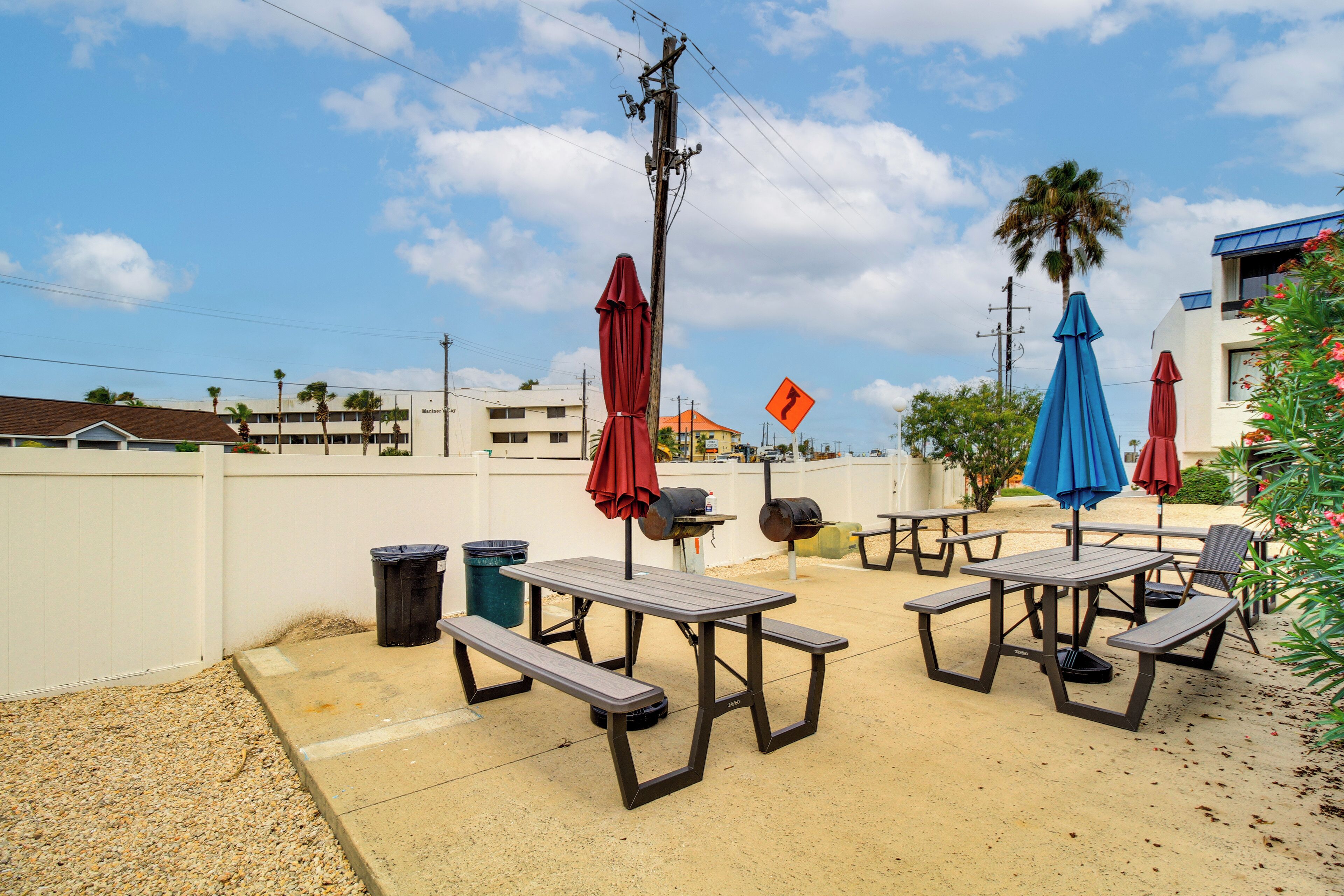 Un espace extérieur aménagé en terrasse, avec des tables de pique-nique et des parasols, entouré d’une clôture blanche et de palmiers.