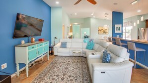 A living room with a white sofa, a blue wall, a ceiling fan, and a TV mounted on the wall. - 3 Mi to Dtwn Fort Myers: Condo w/ Pool & Hot Tub (North Fort Myers)