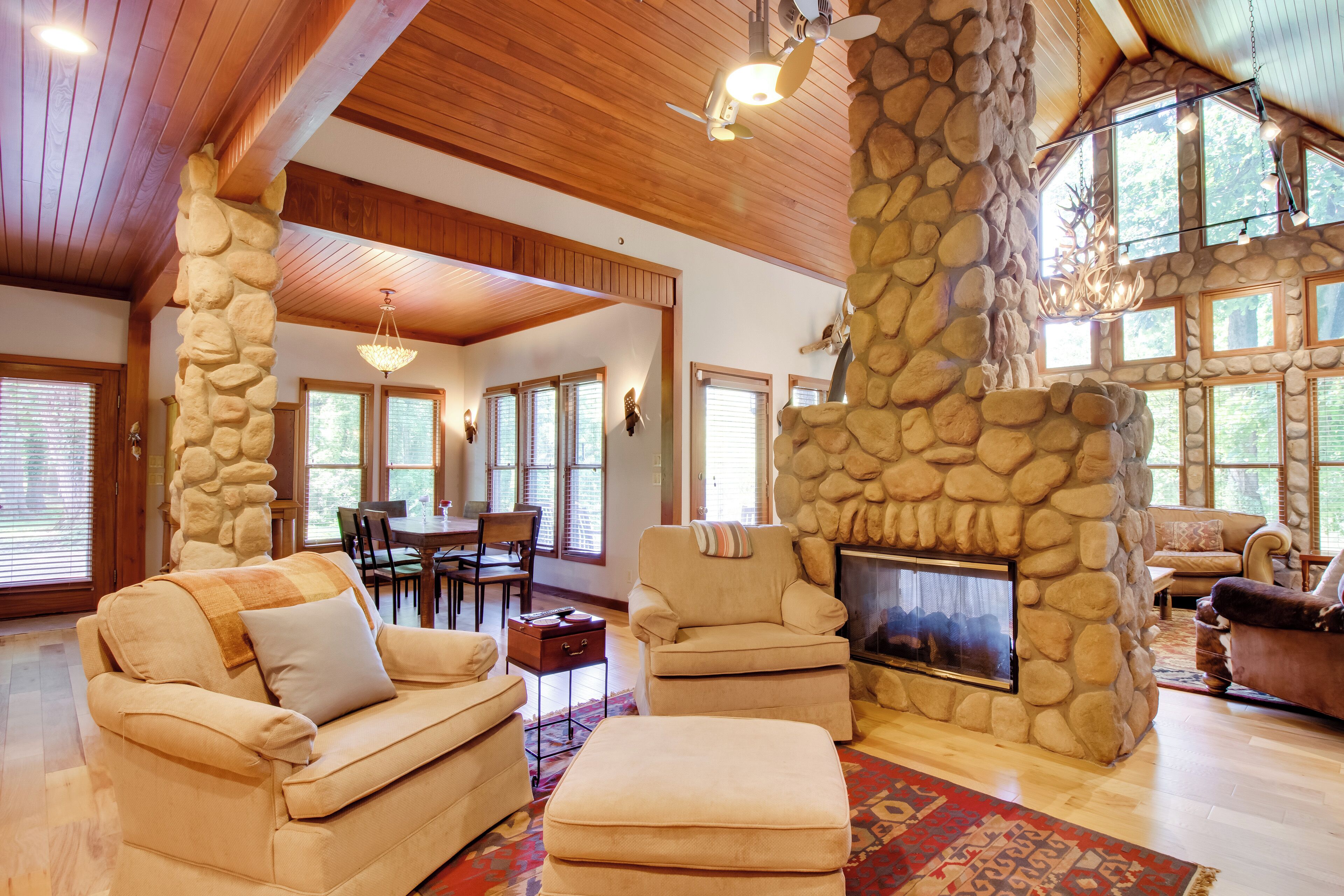A spacious living room with a stone fireplace, wooden ceiling beams, and large windows.