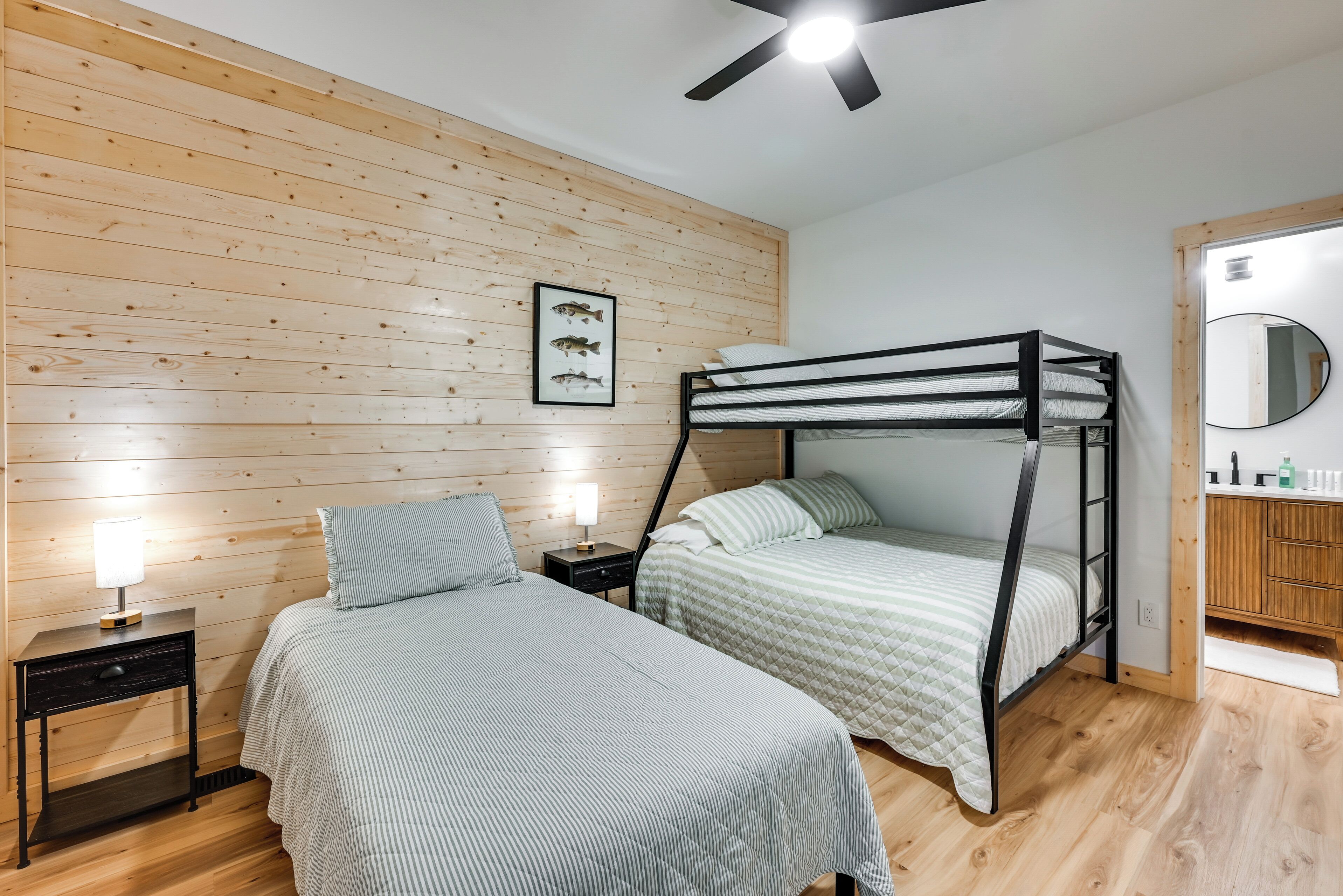 A bedroom with two beds, wooden paneling on the walls, a ceiling fan, and a small bedside table with a lamp.