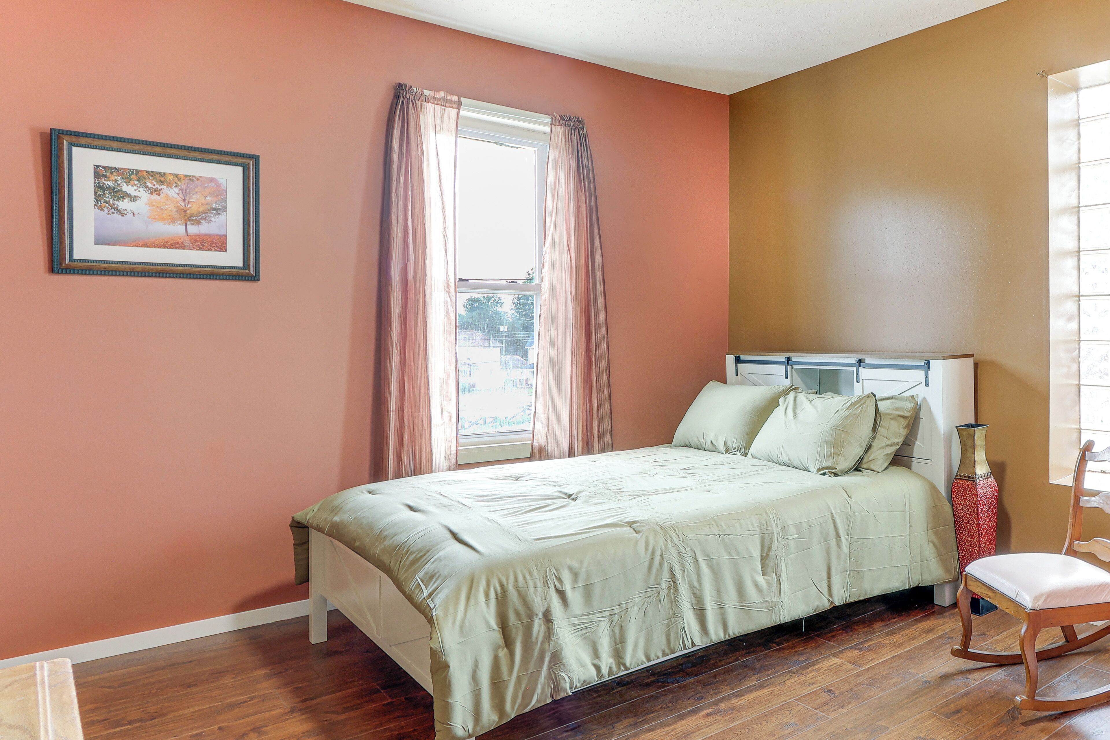 A bedroom with a bed, a chair, a window with curtains, and a framed picture on the wall.