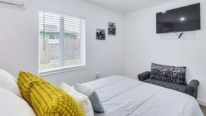 Un dormitorio con una cama, un sofá gris, un televisor montado en la pared y una ventana con persianas.