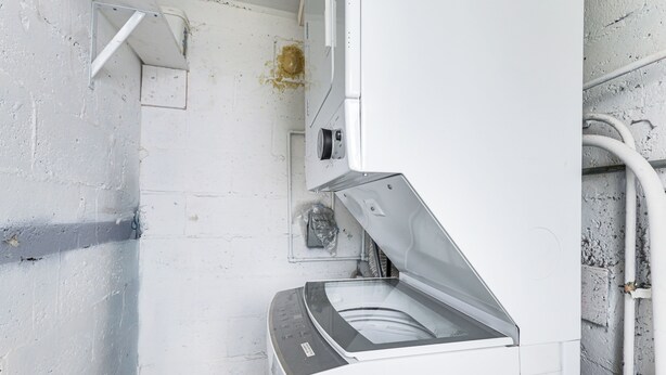 Una lavadora blanca y compacta en una habitación con pared blanca y tuberías a la vista.