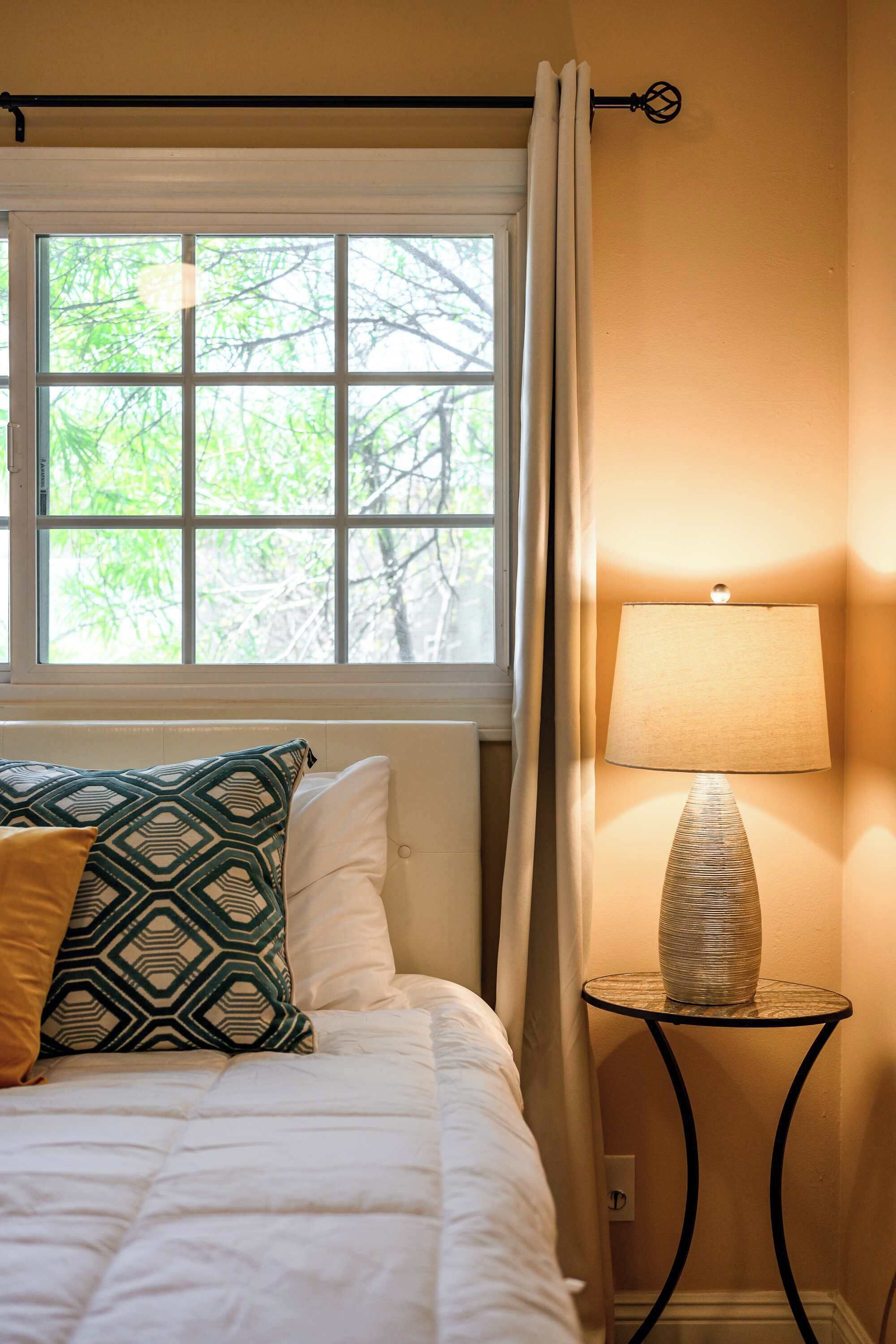 A bedroom with a bed, a window with curtains, a bedside lamp, and a small table.