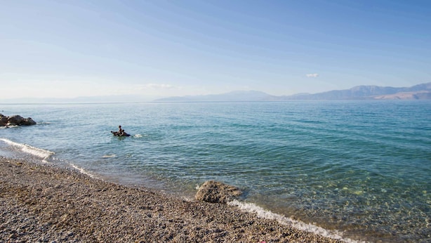 Een kiezelstrand met helder water en verre bergen.