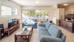 Een ruime woonkamer met een grote grijze bank, een salontafel en een televisie op een houten standaard. Er is een balkon met rieten meubilair en uitzicht op bomen.