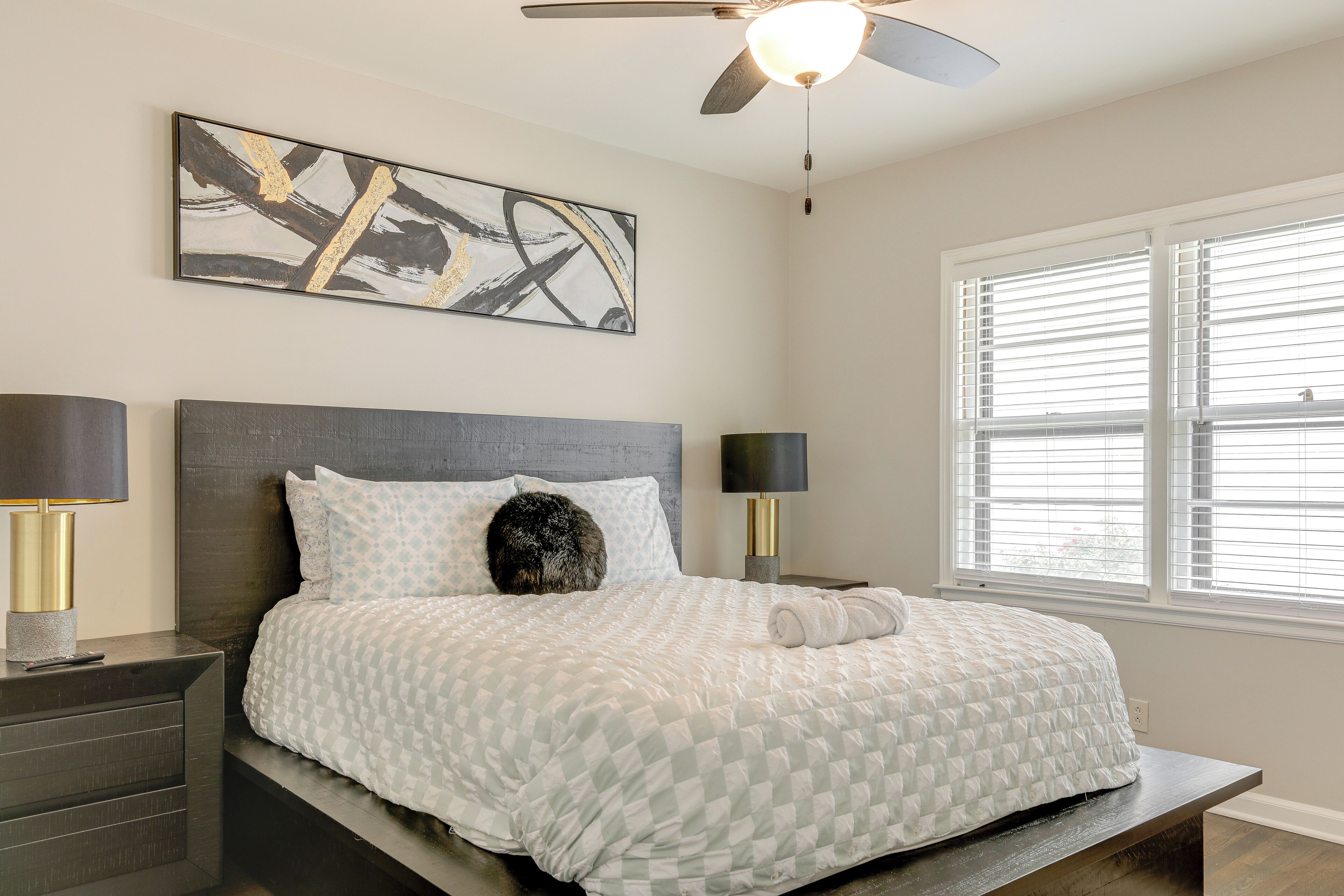 A bedroom with a bed, two bedside tables with lamps, a ceiling fan, a window with blinds, and a wall-mounted abstract painting.