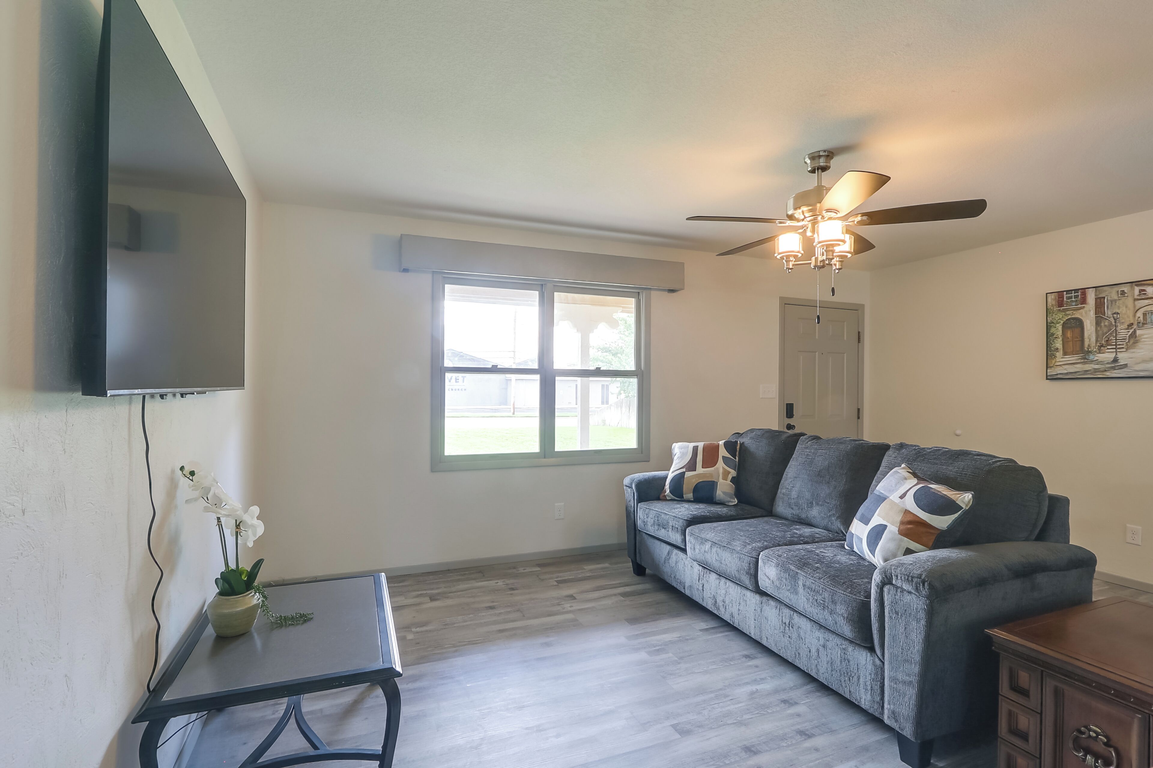 A living room with a gray sofa, a small table, a ceiling fan, and a window.