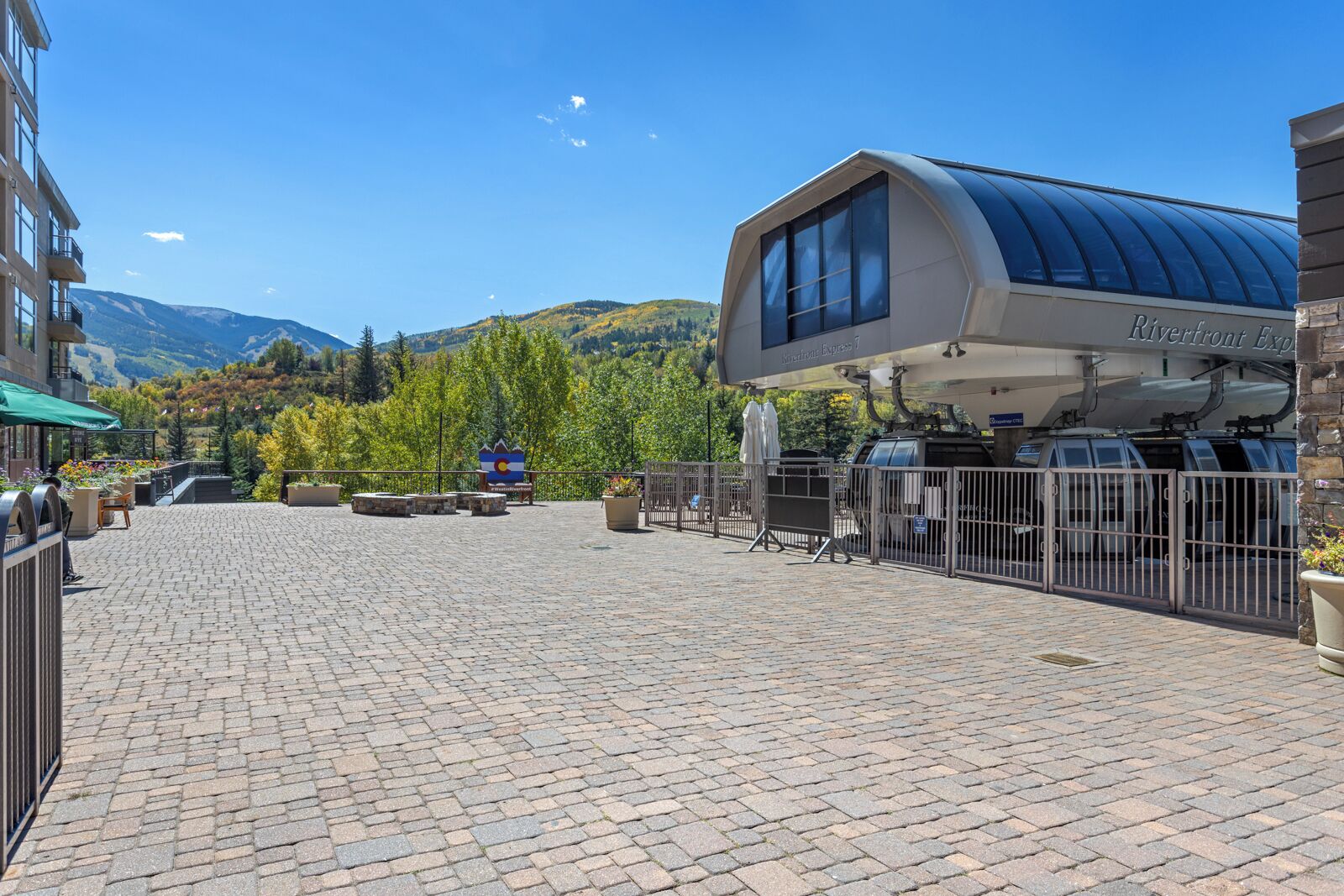 Un espace extérieur aménagé avec une surface pavée, un abribus couvert, un paysage montagneux en arrière-plan et un bâtiment portant une enseigne indiquant « Riverfront Express 7 ».