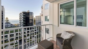 Un balcon avec un fauteuil en osier et un coussin, d’où l’on peut admirer un paysage urbain avec des bâtiments modernes et une vue sur la mer.