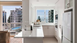 Une cuisine moderne dotée d’une grande fenêtre, de meubles blancs et offrant une vue sur une piscine et des immeubles.