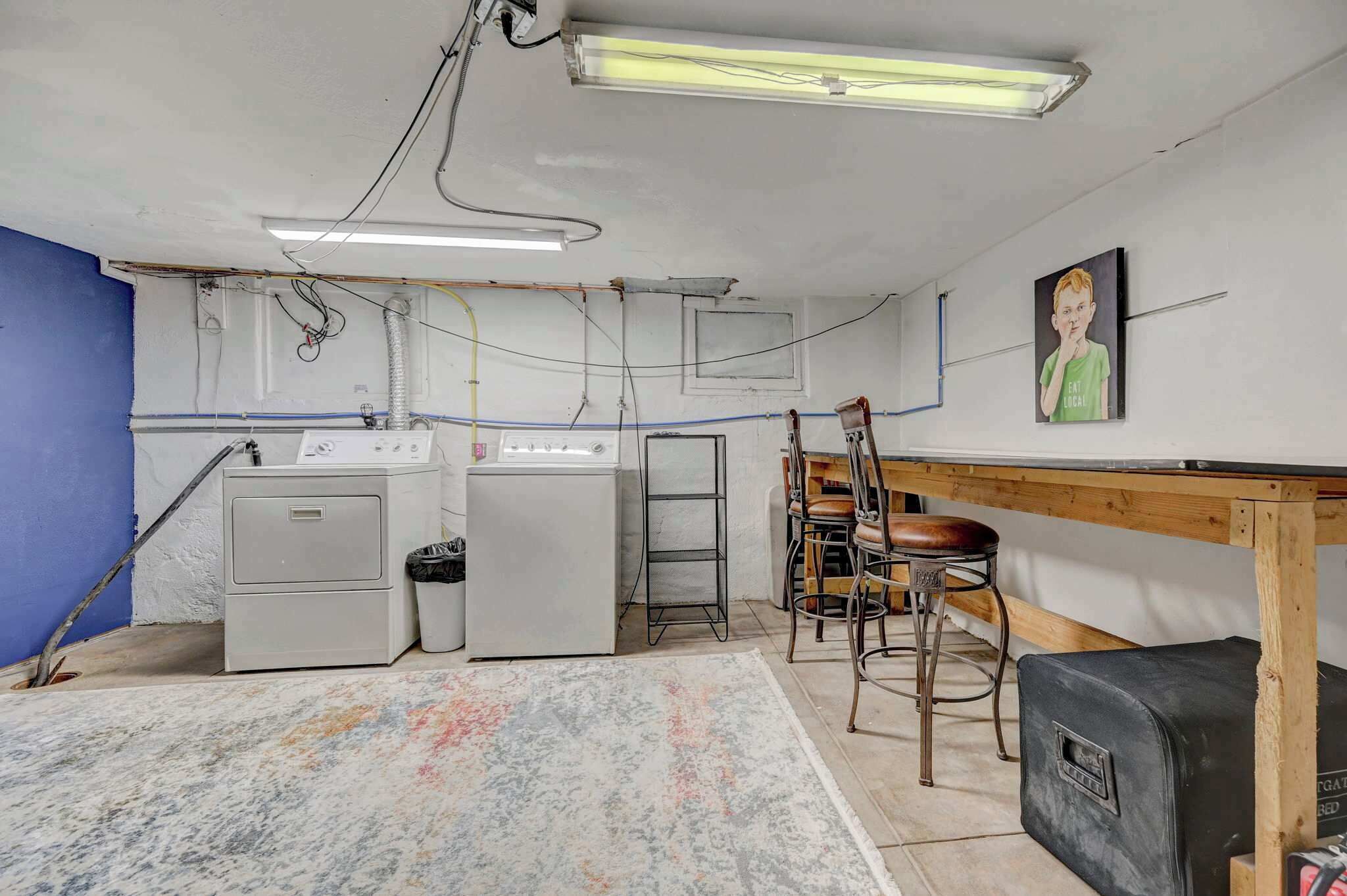A kitchen with a washing machine, a refrigerator, a wooden table with chairs, and a painting on the wall.