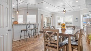 A spacious dining area with a wooden table and chairs, a kitchen with white cabinetry, and a ceiling with exposed beams.