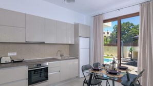 A modern kitchen with a built-in oven, a toaster, and a refrigerator. There is a dining area with a glass table and chairs, and a view of a swimming pool through large windows. - Ninalma Villa (Rhodes)