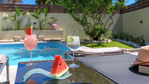 A glass table with two drinks, one with a slice of watermelon, and a hat on the table. - Ninalma Villa (Rhodes)