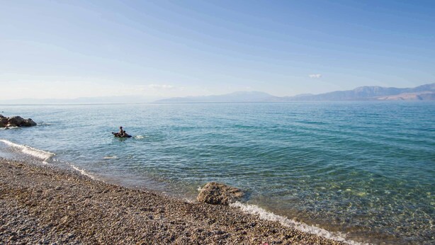 A pebbled beach with clear water, a person in a kayak, and distant mountains.