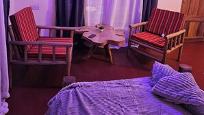 A room with a bed, two striped chairs, a wooden table, and a window with purple curtains.