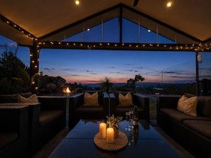 A covered outdoor seating area with a view of the sunset, a table with candles, and a bottle of wine.
