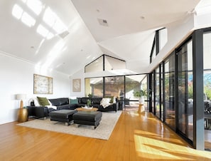A spacious living area with a high ceiling, a black leather sofa set, a coffee table, and a large sliding glass door leading to an outdoor patio.