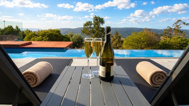 Un salon de bord de piscine avec une bouteille de vin et des verres, offrant une vue sur un ciel bleu dégagé et un paysage montagneux.