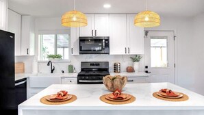 Una cocina moderna con gabinetes blancos, un microondas negro y una estufa. Hay dos lámparas colgantes y una isla grande con un bol y platos.
