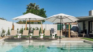 Una piscina con sillas de descanso y sombrillas, rodeada por un área cercada con vegetación.