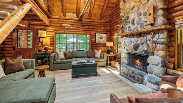 A log cabin interior with a stone fireplace, wooden beams, and a cozy seating area.