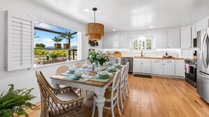 A modern kitchen with white cabinetry, a dining area with a wooden table and wicker chairs, and a view of greenery outside.