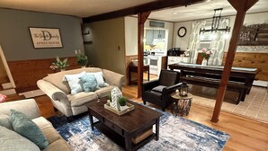 A living room with a sofa, coffee table, and a rug. There is a kitchen in the background with cabinets and a dining table. - Spacious 6-bedroom home just steps from outdoor adventures! (Laconia)