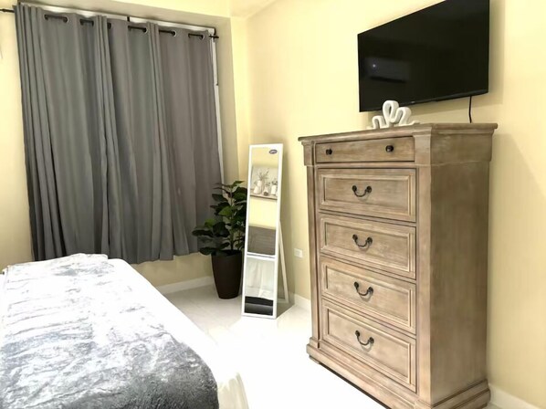 A bedroom with a bed, a dresser, a potted plant, a mirror, and a TV mounted on the wall. - Stylish 1-Bed Condo with Views in Harloe (Kingston)