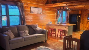 Un acogedor interior de cabaña con un sofá, un tapete estampado, una zona de comedor con taburetes y una cocina con refrigerador.