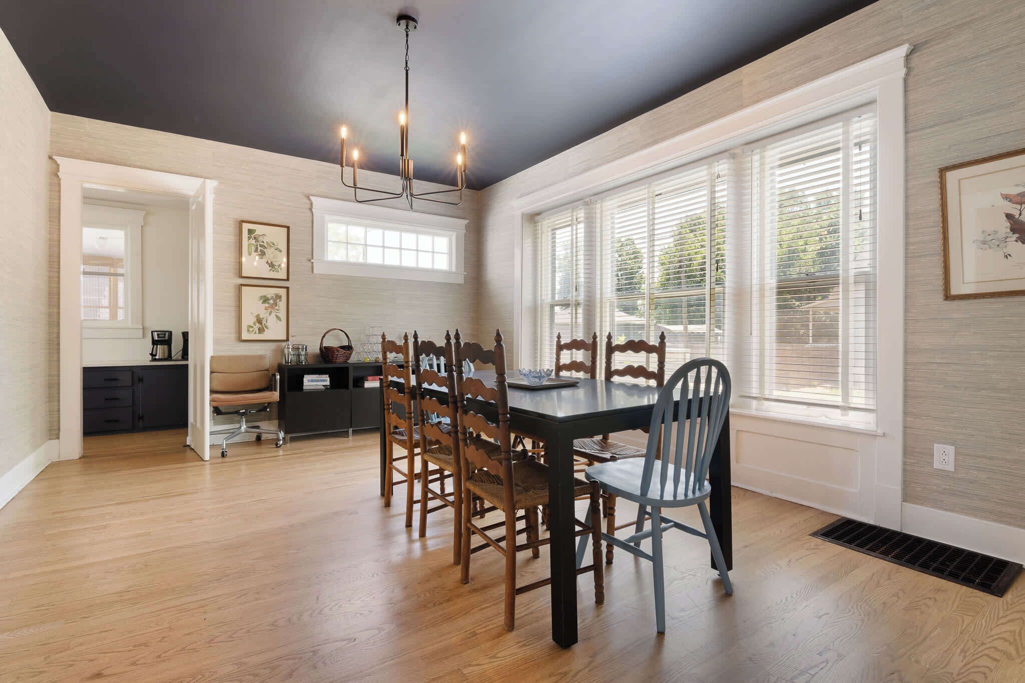 A dining area with a dark ceiling, wooden dining table, and chairs. There are framed pictures on the walls and a window with blinds.