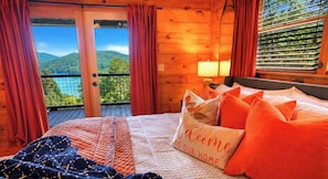 A wooden cabin room with a bed, a welcome pillow, a view of mountains, and a balcony.