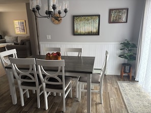 Zona pranzo con tavolo in legno, sedie bianche, un lampadario e due quadri appesi al muro.