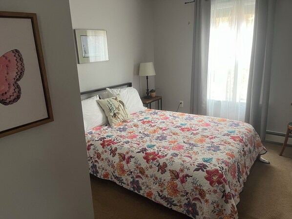 Un dormitorio con cubrecama floral, una lámpara de noche, un cuadro enmarcado en la pared y una ventana con cortinas.