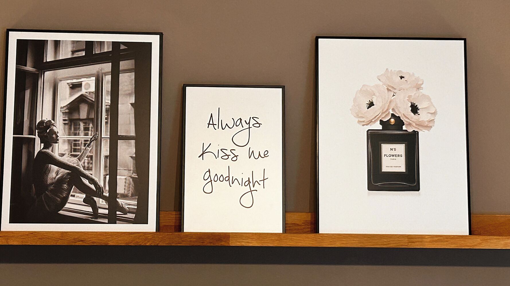Drei gerahmte Objekte auf einem Regal: ein Schwarz-Weiß-Foto einer Person vor einem Fenster, ein gerahmter Text mit der Aufschrift 'Küss mich immer zur Nacht', und eine gerahmte Parfumflasche mit dem Aufdruck 'N'S FLOWERS'.
