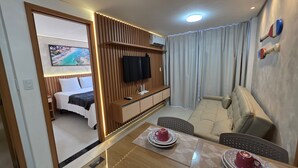 A hotel room with a bed, a TV, a sofa, a dining table with a red cup, and a view of a beach through the window.