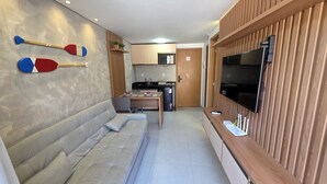 A modern living room with a sofa, a dining table, a kitchen area, and a TV mounted on the wall.
