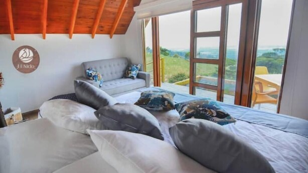 A bedroom with a large bed, a grey sofa, and a view of the outdoors through a large window.