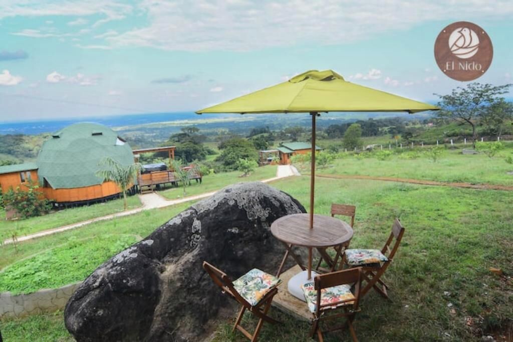 An outdoor seating area with a table and chairs, a large rock, and a view of a green landscape with buildings in the background.