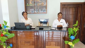 A reception desk with two staff members, a laptop, a small screen, and a decorative plant. - Hotel Indigo (Kinshasa)