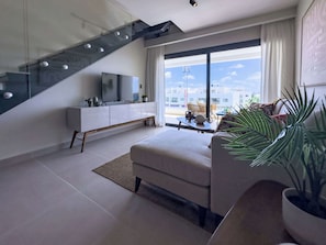 A modern living room with a glass staircase, a flat-screen TV, a sofa, and a view of the cityscape.