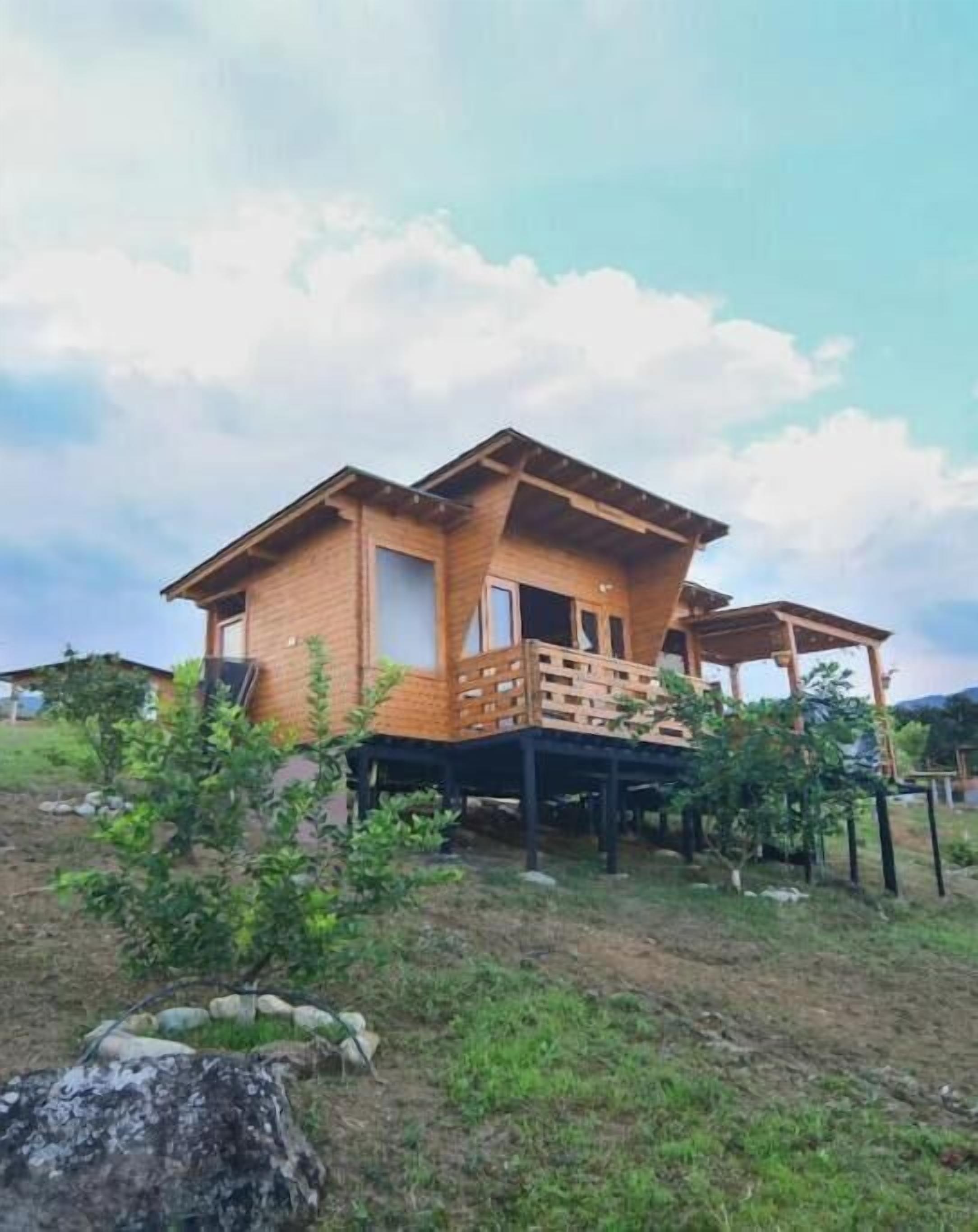 A wooden house with a balcony and a small deck, surrounded by greenery and a clear sky.