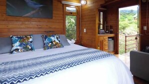 A wooden cabin bedroom with a bed, pillows, a painting, a window, and a view of greenery. - Romantic Cabin with Jacuzzi Azulejo (Acacías)