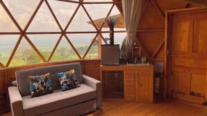 Una habitación acogedora con un gran ventanal que ofrece vistas al campo, un escritorio de madera con una computadora portátil y un sofá con cojines estampados.