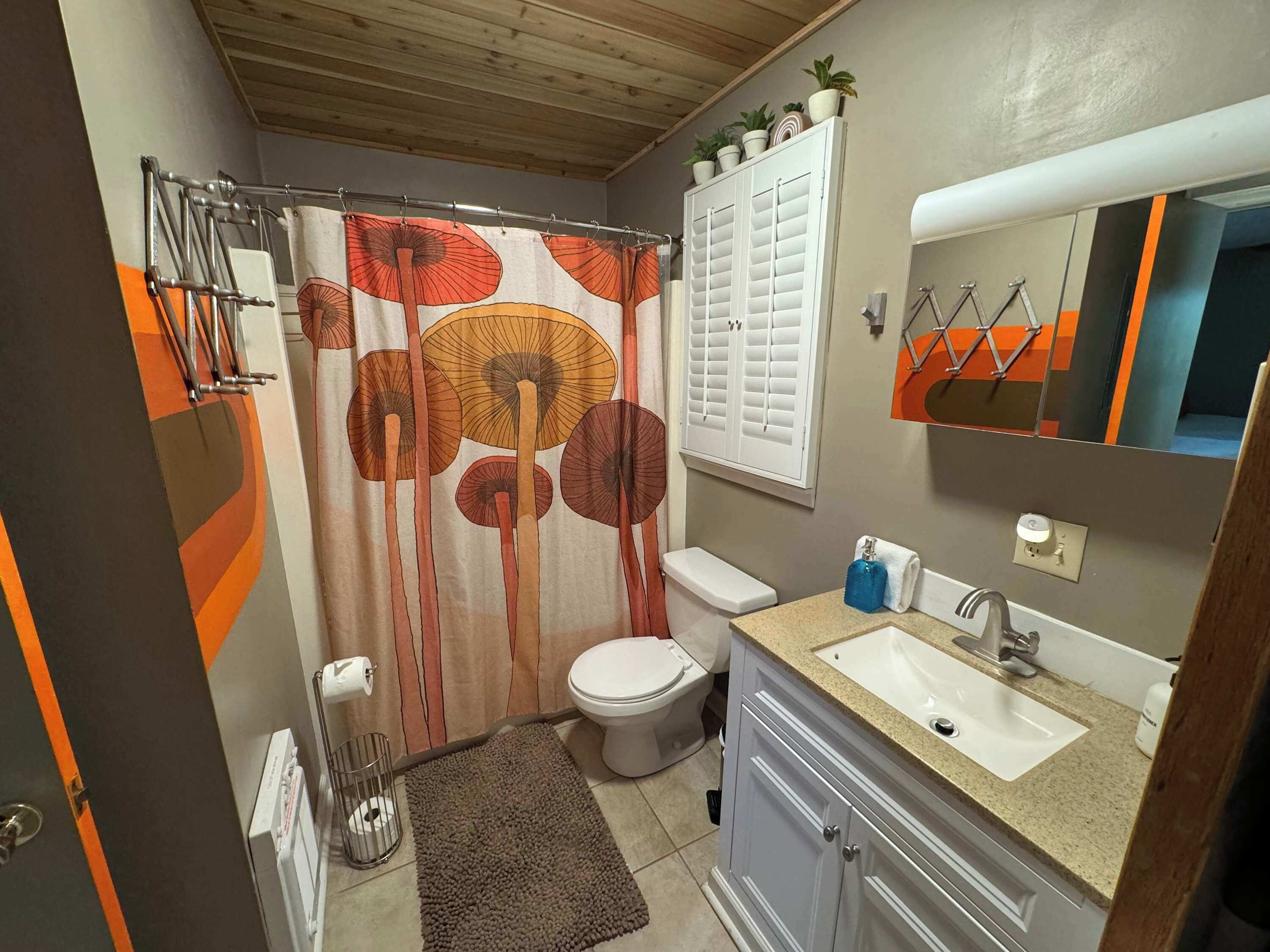 Ein Badezimmer mit Duschvorhang mit Pilzmuster, Toilette, Waschbecken mit Spiegel und kleinem Fenster mit Rollläden.