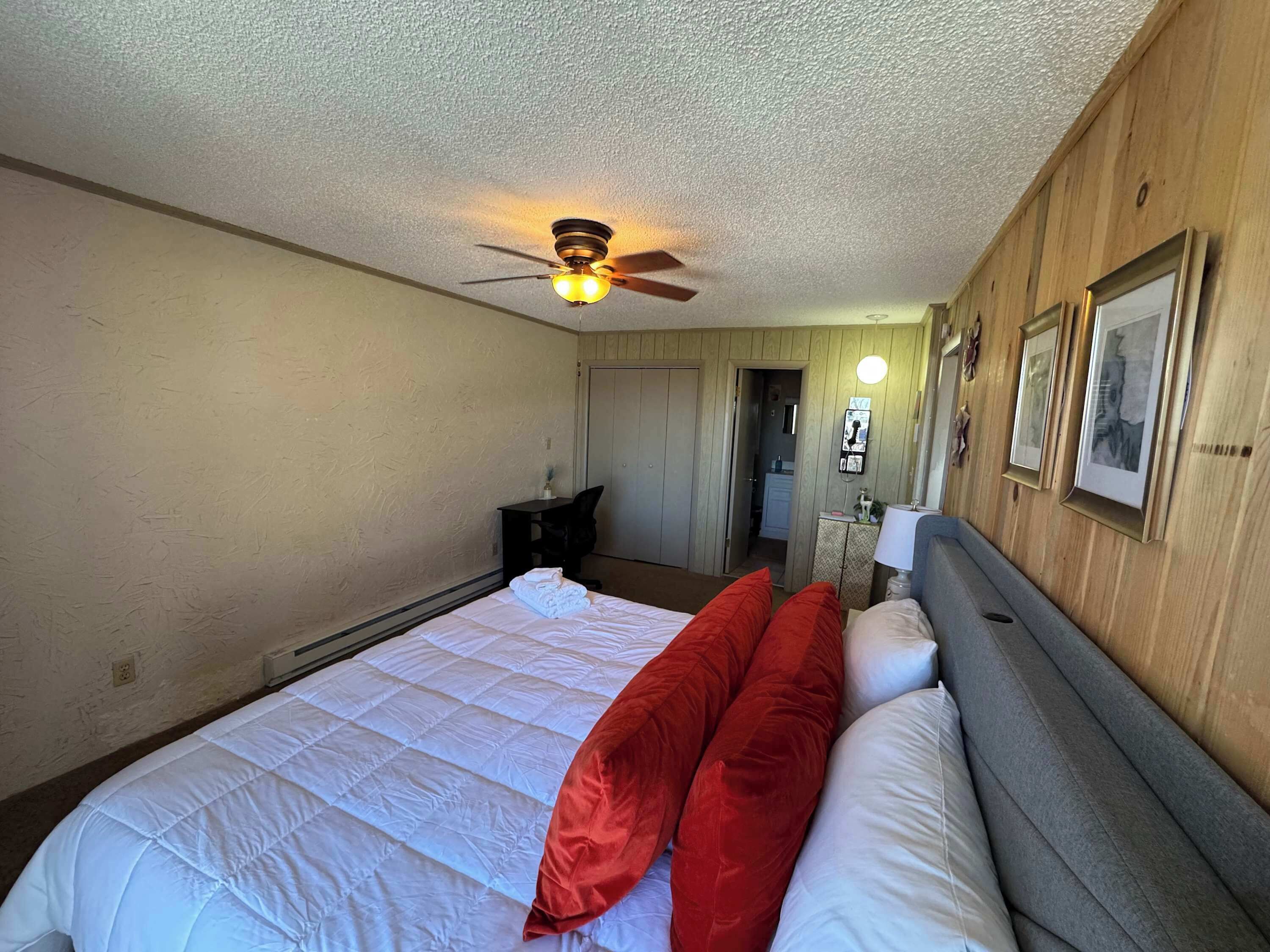 A bedroom with a large bed, a ceiling fan, wooden walls, and a door leading to a bathroom.