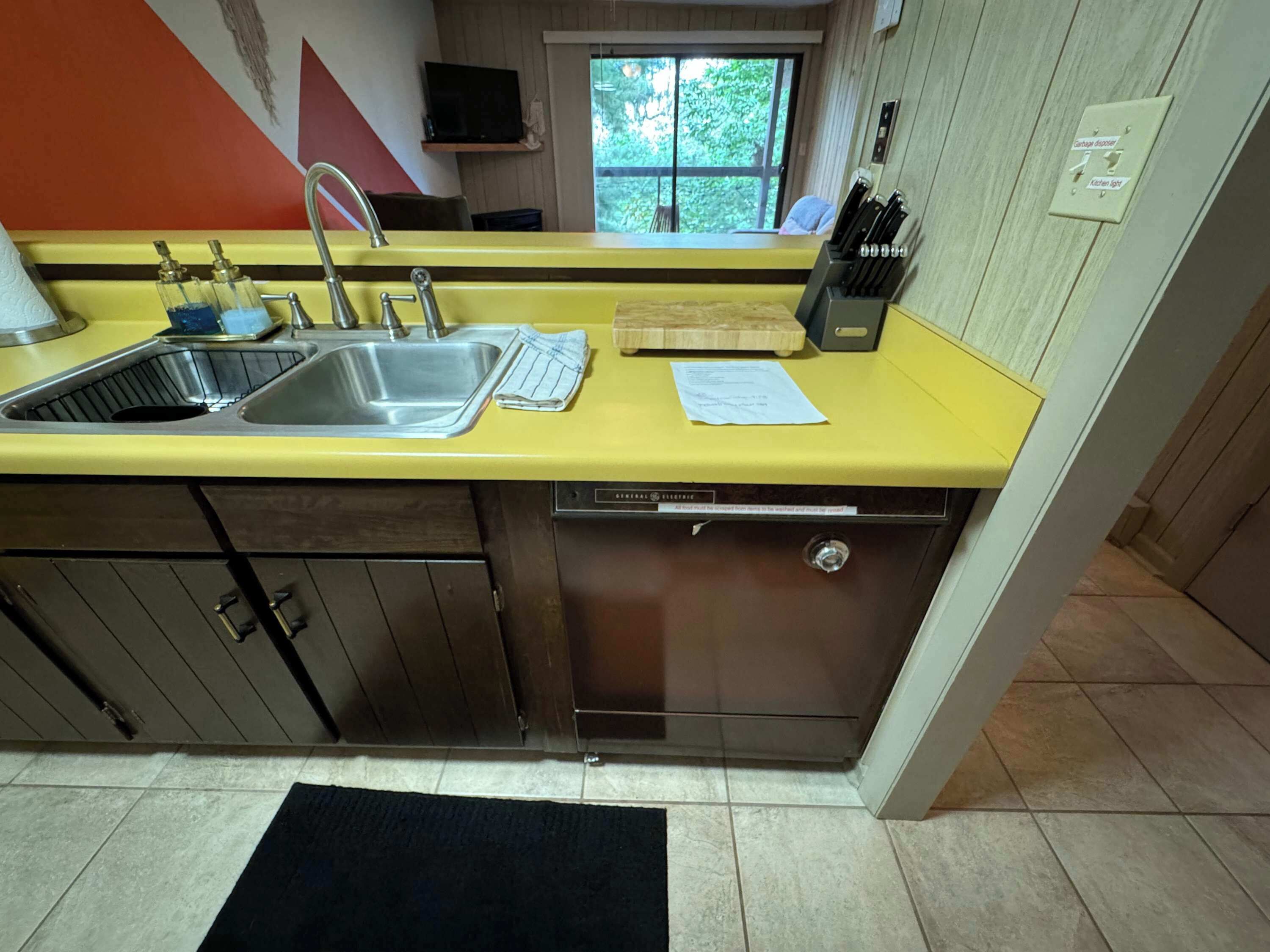 A kitchen with a yellow countertop, stainless steel sink, and a dishwasher. There is a window with a view of greenery outside.
