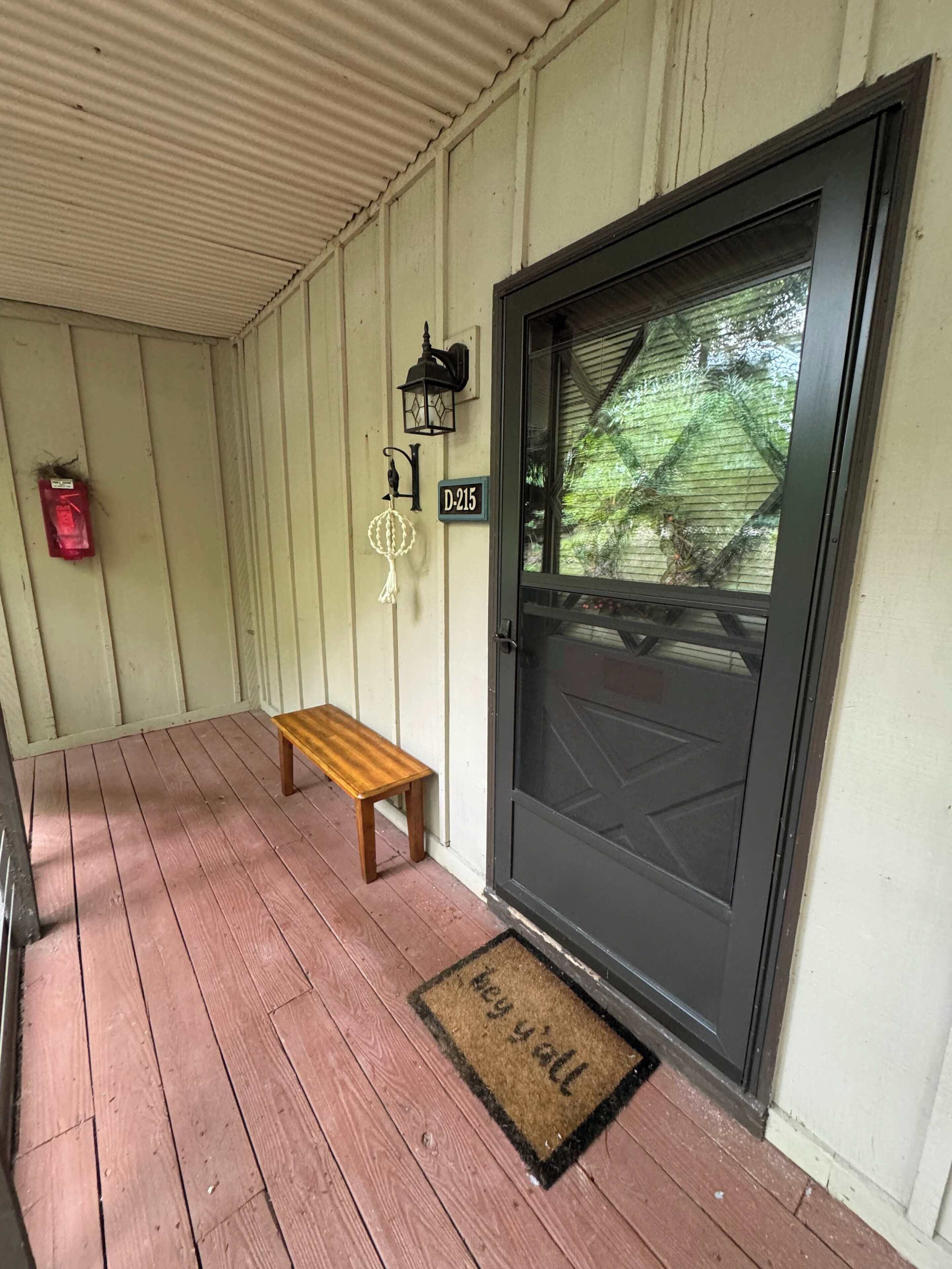 A wooden porch with a bench, a black door with a glass panel, a lantern, and a mailbox.