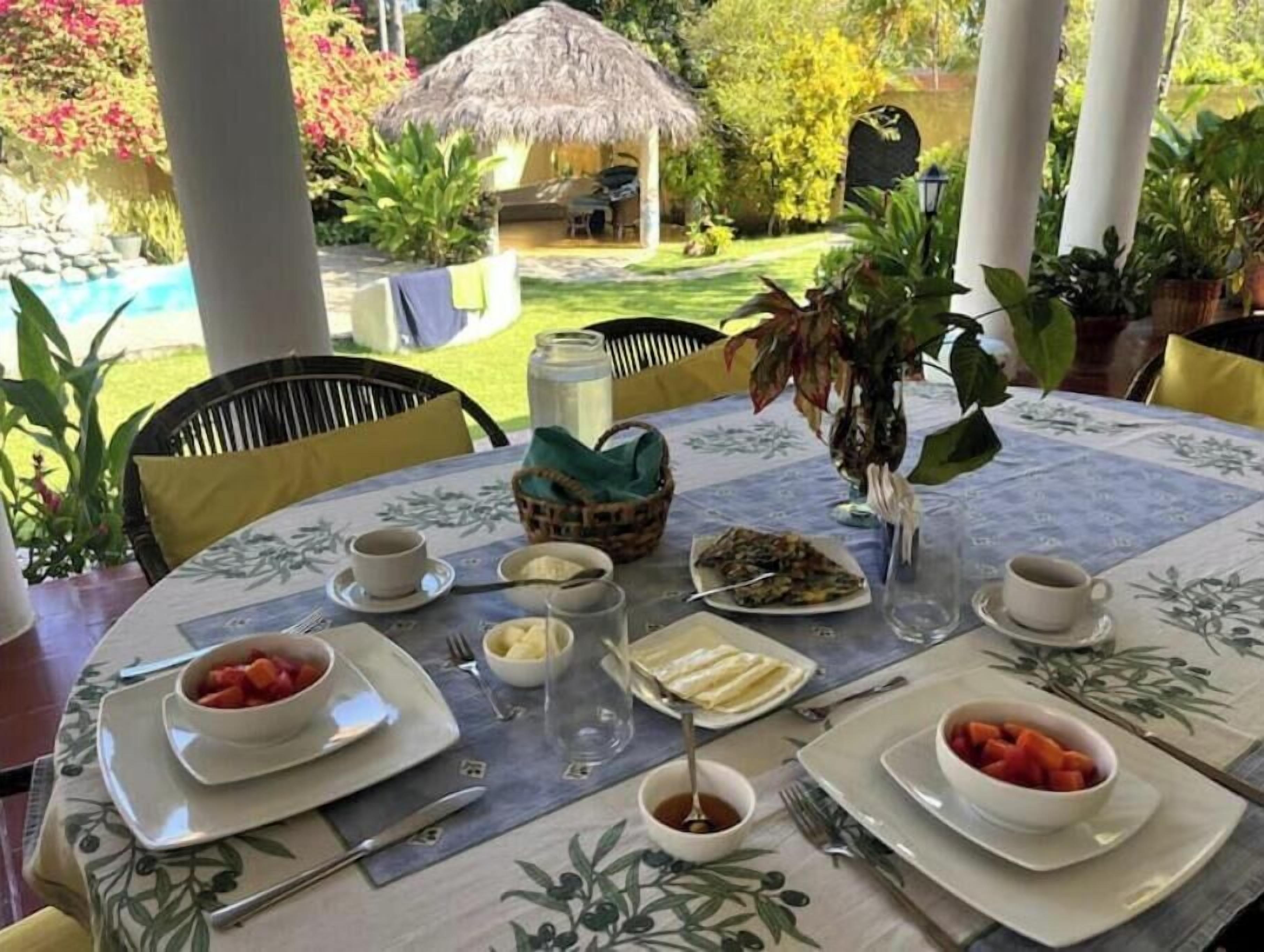 A dining table set with plates of food, cups, and cutlery, surrounded by chairs, with a thatched-roof structure and lush greenery in the background.